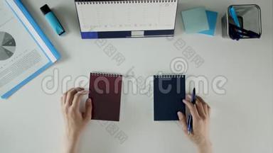 一个男人坐在他的办公桌前，拿着两本笔记本。 桌子上有办公用品。